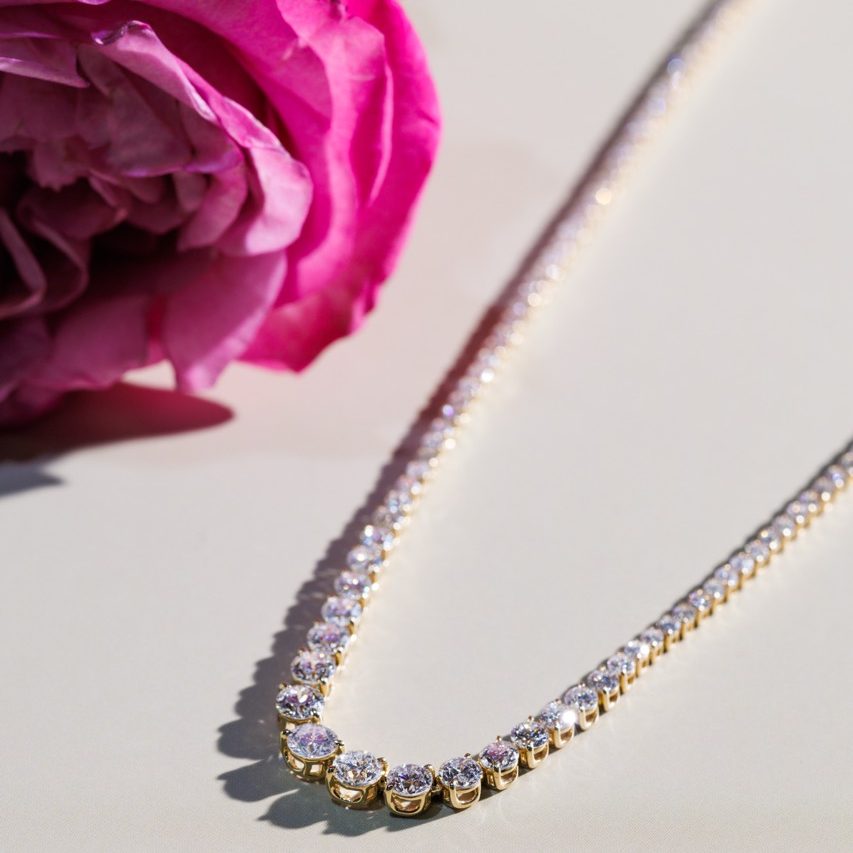 Elegant gold diamond tennis necklace displayed on a light surface beside a blooming pink rose, showcasing fine craftsmanship and luxury design