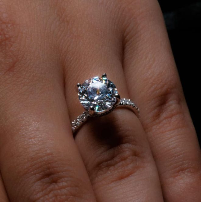 Close-up of a solitaire engagement ring with a round brilliant-cut diamond on a woman's finger, featuring a diamond-paved band and prong setting